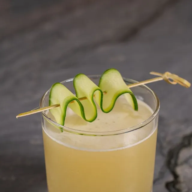A pale gin cocktail on ice with a ribbon of cucumber on a pick.