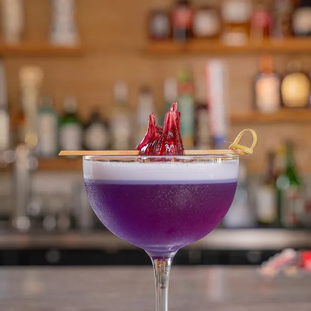 A purple cocktail with a white foam top, garnished with a dried hibiscus flower.