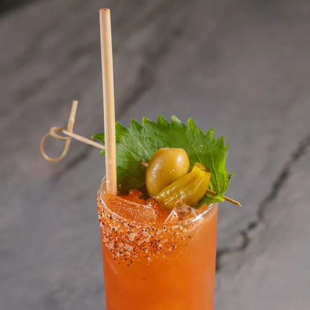 A red-orange cocktail with a chili-salt rim, garnished with a shiso leaf and peppers.